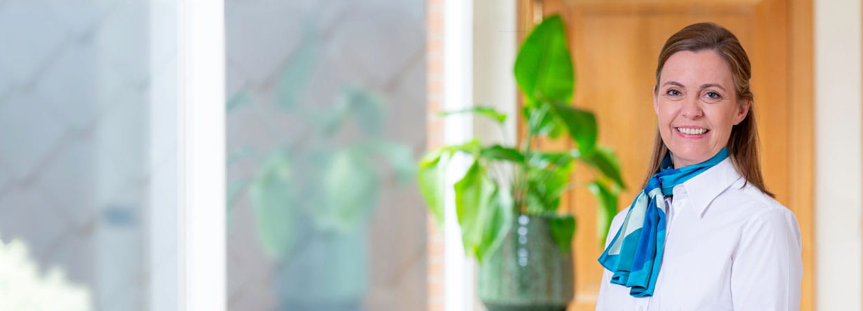 Persoon in witte blouse met blauwe sjaal staat binnen in een lichte ruimte, met een groene plant en houten wand op de achtergrond.